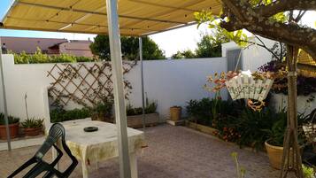 Een overdekt terras met een tafel en stoelen, omringd door potplanten en een decoratieve ventilator.