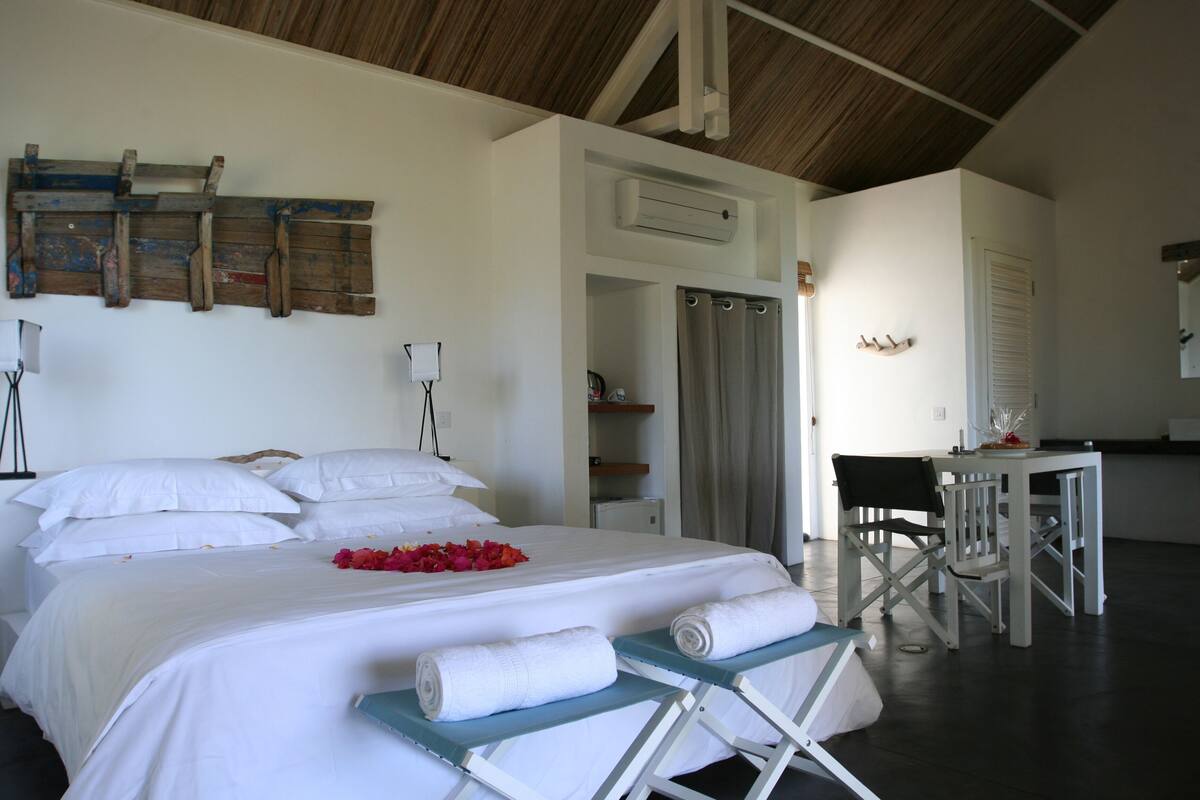 A hotel room with a large bed, a desk, and a chair. There is a shower area and a door leading to another room.