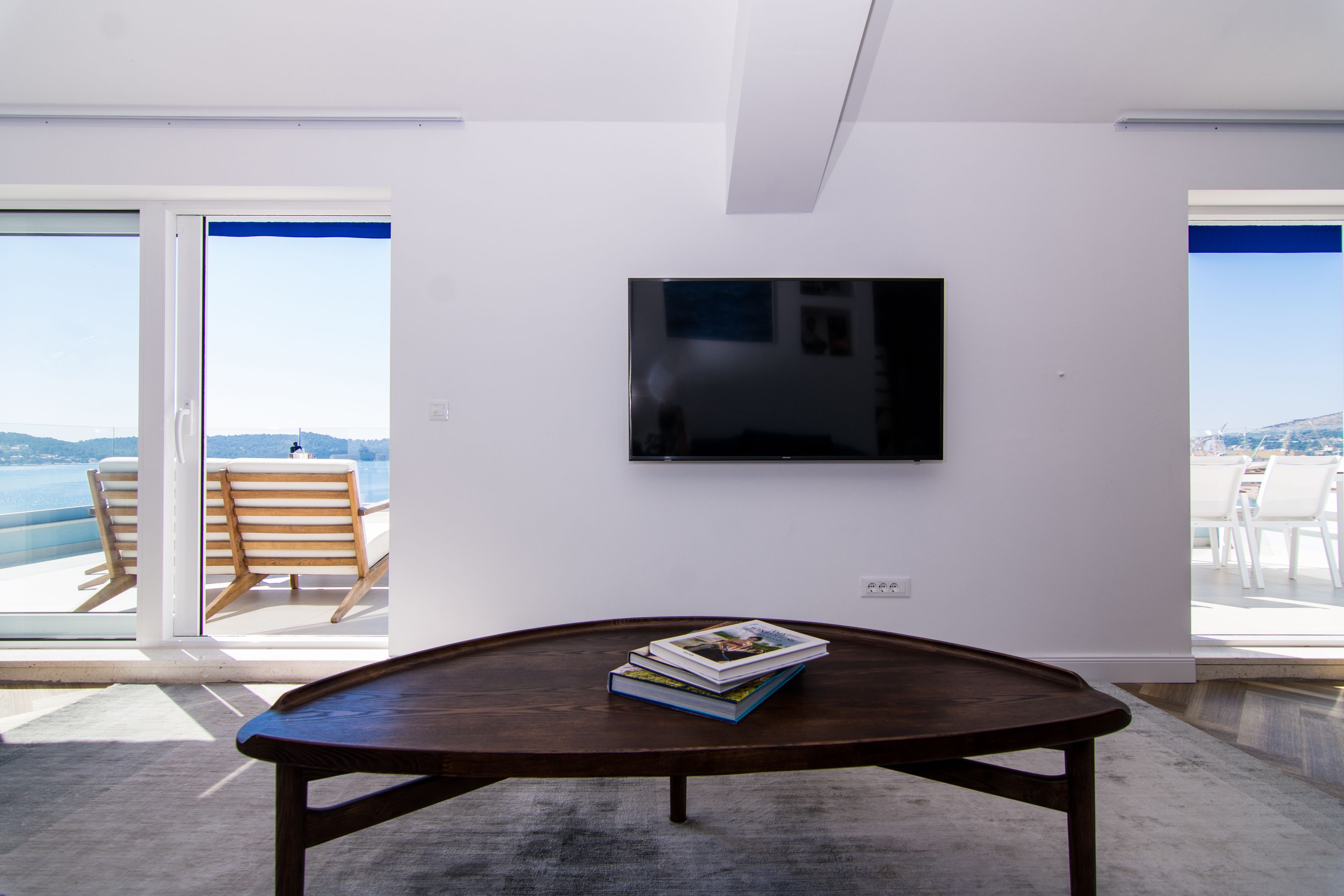 A modern living room with a large flat-screen TV mounted on the wall, a wooden coffee table with books, and a view of the ocean through large sliding glass doors.