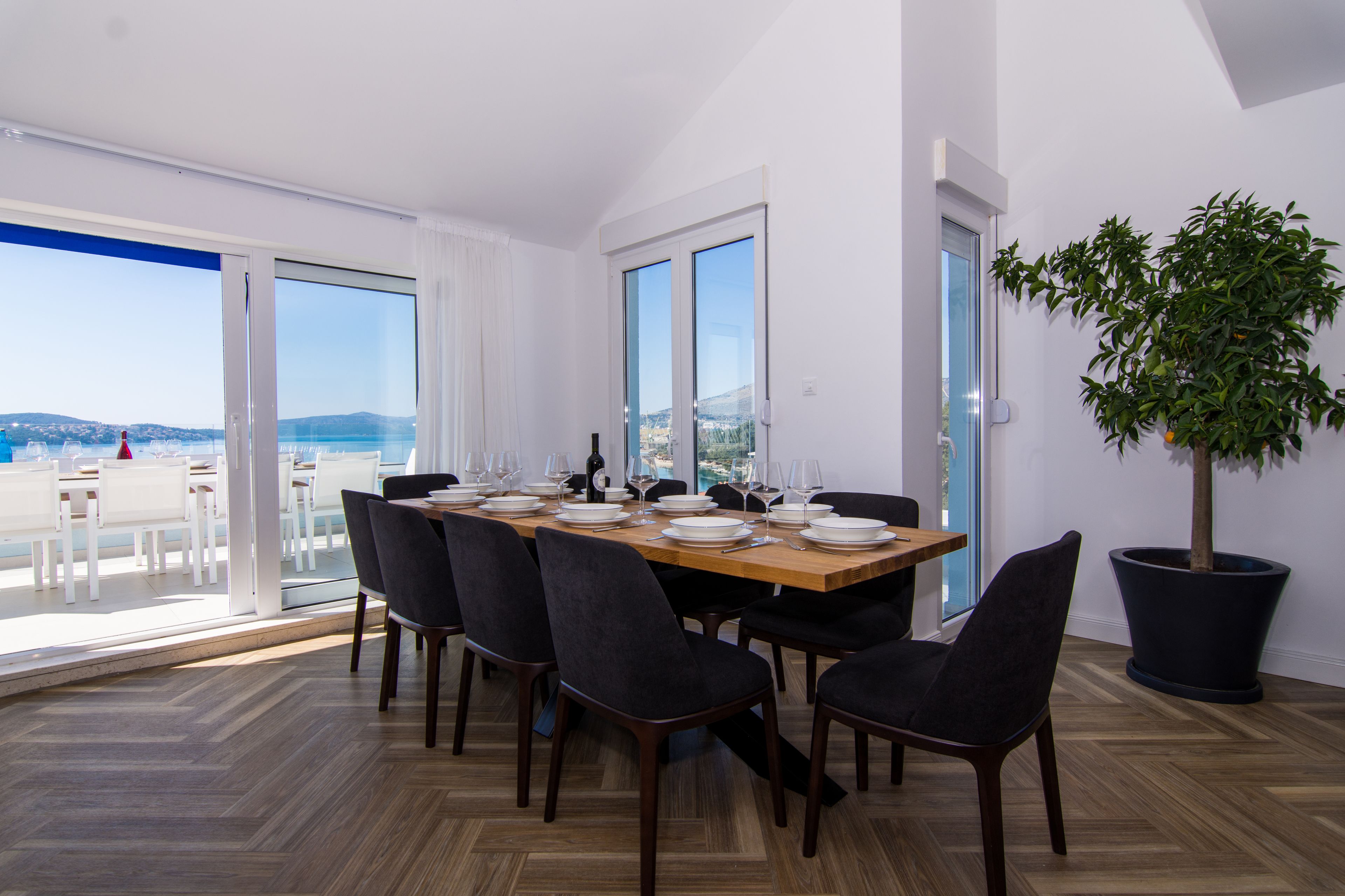 A dining area with a large table set for six, large windows, and a potted plant.