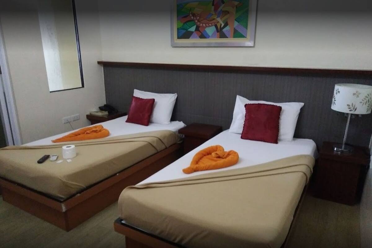 Two single beds with beige bedding, red pillows, and orange towels. There is a nightstand with a lamp and a phone on each bed. A painting is on the wall above the beds.