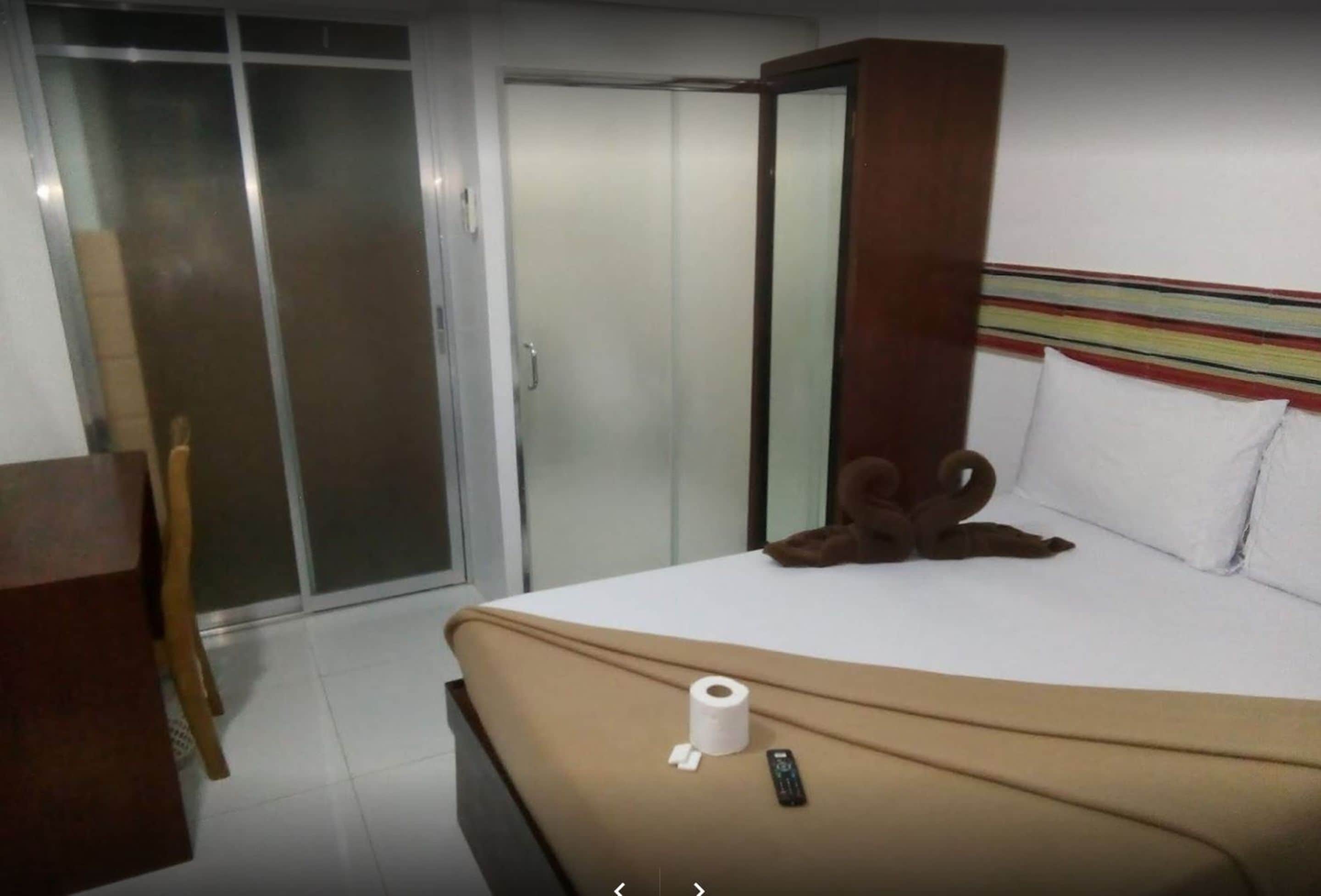 A hotel room with a bed, a wooden headboard, a pair of slippers, a remote control, and a toilet paper roll on the bed.