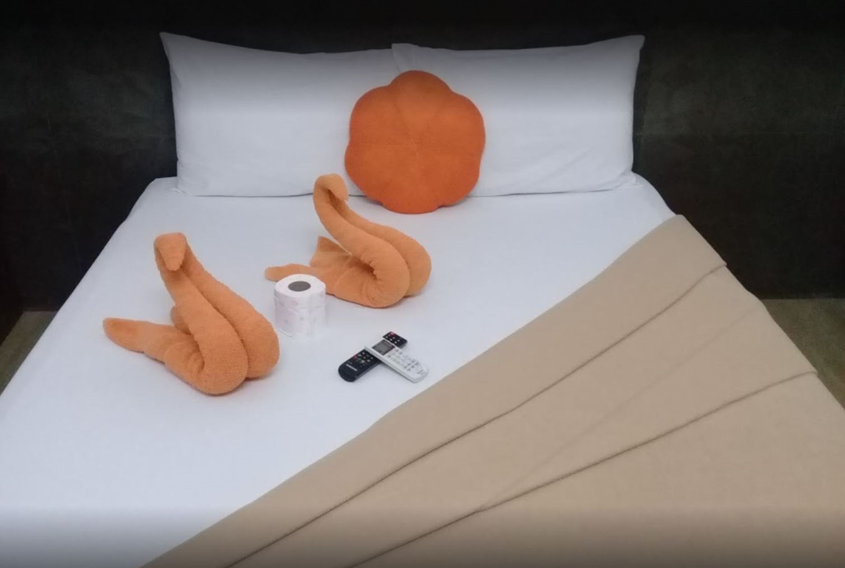 A bed with a white sheet, an orange pillow, a beige blanket, and a remote control.