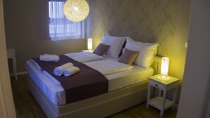 Una cama bien hecha con sábanas blancas y un plaid marrón, una mesita de noche con una lámpara, y una pared con estampado.