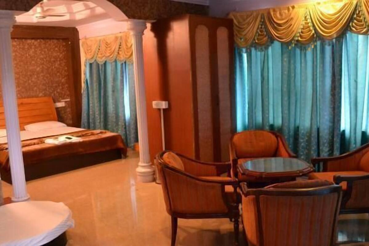 Une chambre à coucher avec un lit à baldaquin, un coin salon avec deux fauteuils et une petite table, et une grande fenêtre avec des rideaux.