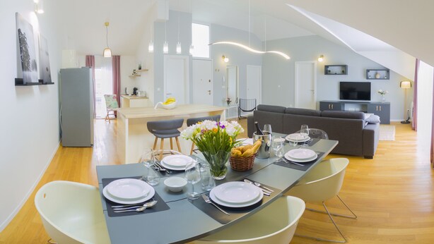 A modern dining area with a long table set for four, a kitchen in the background, and a living area with a sofa and TV. - Presidential Suites Chica Cure (Split)