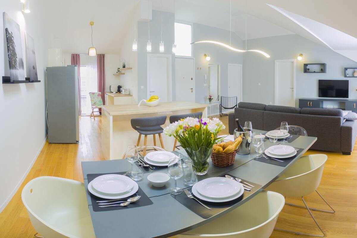 A modern dining area with a long table set for four, a kitchen in the background, and a living area with a sofa and TV.
