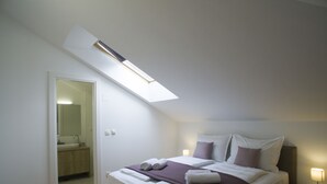 A bedroom with a skylight, a bed with pillows and a blanket, a bedside table with a lamp, and a bathroom visible through an open door. - Presidential Suites Chica Cure (Split)