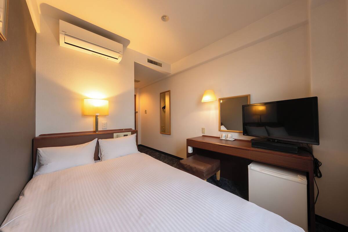 Una habitación de hotel con cama, lámparas de noche, televisor, escritorio y un refrigerador pequeño.