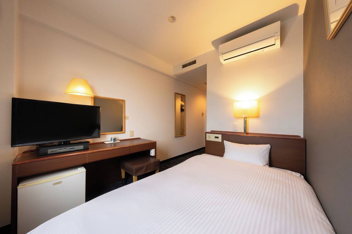 Una habitación de hotel con cama, televisión, escritorio con silla y un refrigerador pequeño.