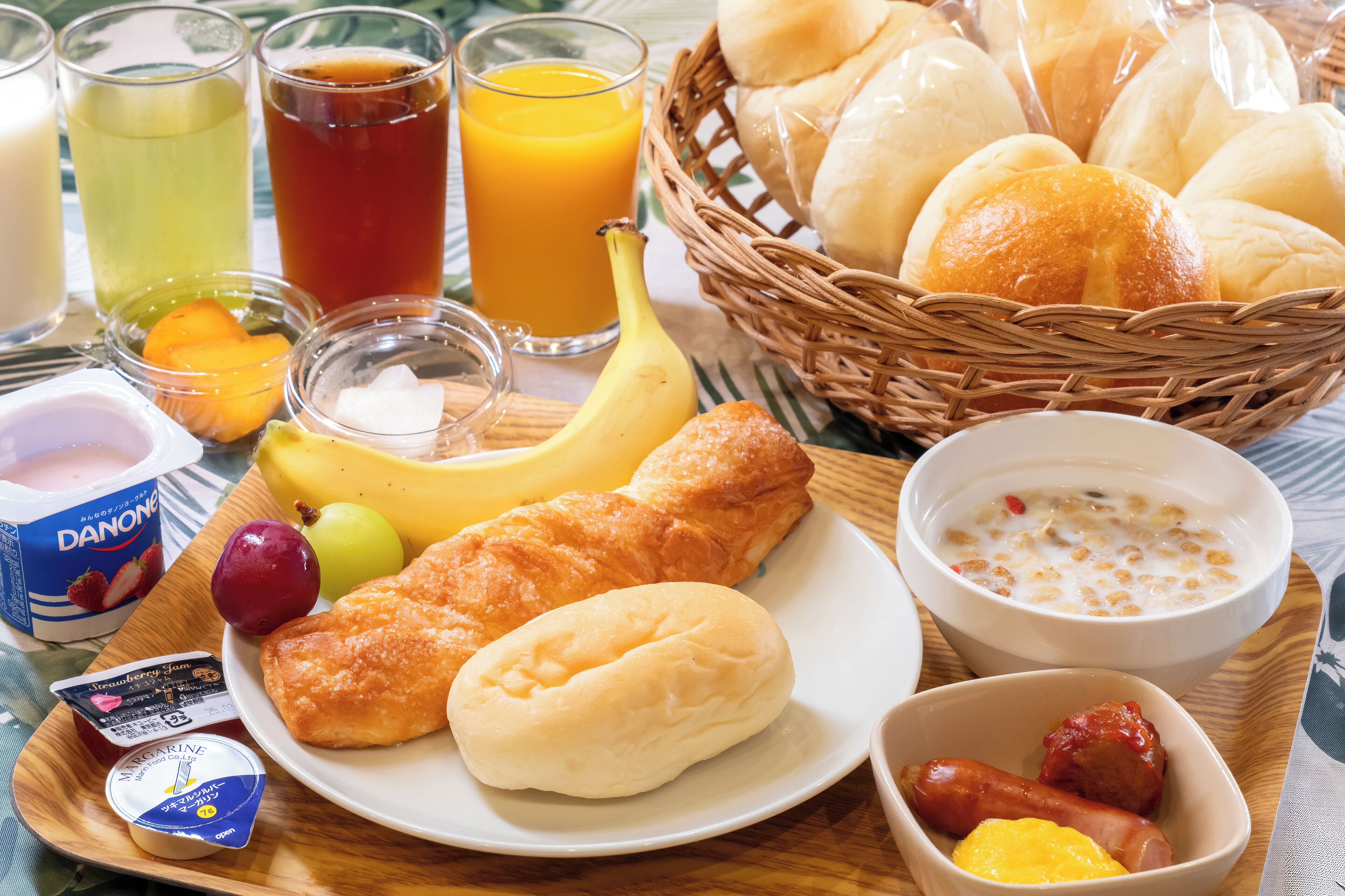 Bandeja de desayuno con diversos alimentos, incluyendo pan, un plátano, un tazón de sopa y un plato con salchichas.