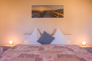 A bedroom with a bed, two bedside lamps, a floral quilt, and a sunset painting on the wall. - Relaxing time out in the Silbermöwe vacation apartment on the Baltic Sea! (Dahme)