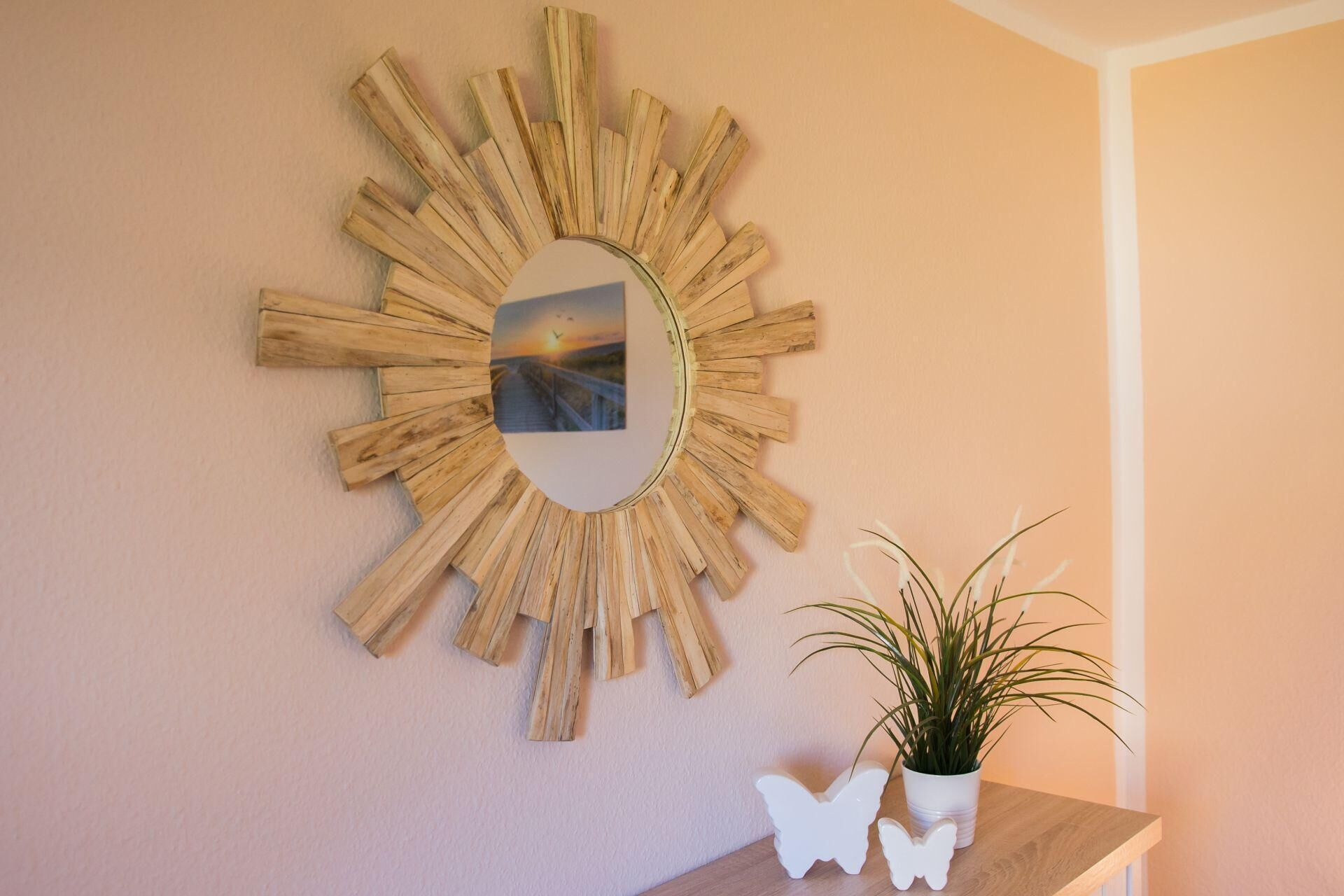 A wall-mounted wooden sunburst mirror with a picture inside, a white potted plant, and two white butterfly-shaped ornaments on a wooden table.