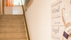 Una escalera de madera que sube a un rellano con un cartel en la pared que dice "Du kannst den Wind nicht ändern, aber die Segel richtig setzen".