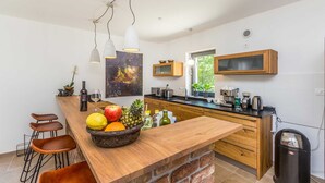A modern kitchen with a wooden island, bar stools, a fruit bowl, and a view of greenery outside. - Holiday house RASOPASNO (Rasopasno)