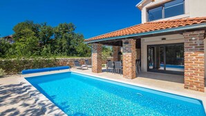 A swimming pool with a covered patio area, stone wall, and a view of a house with a tiled roof. - Holiday house RASOPASNO (Rasopasno)