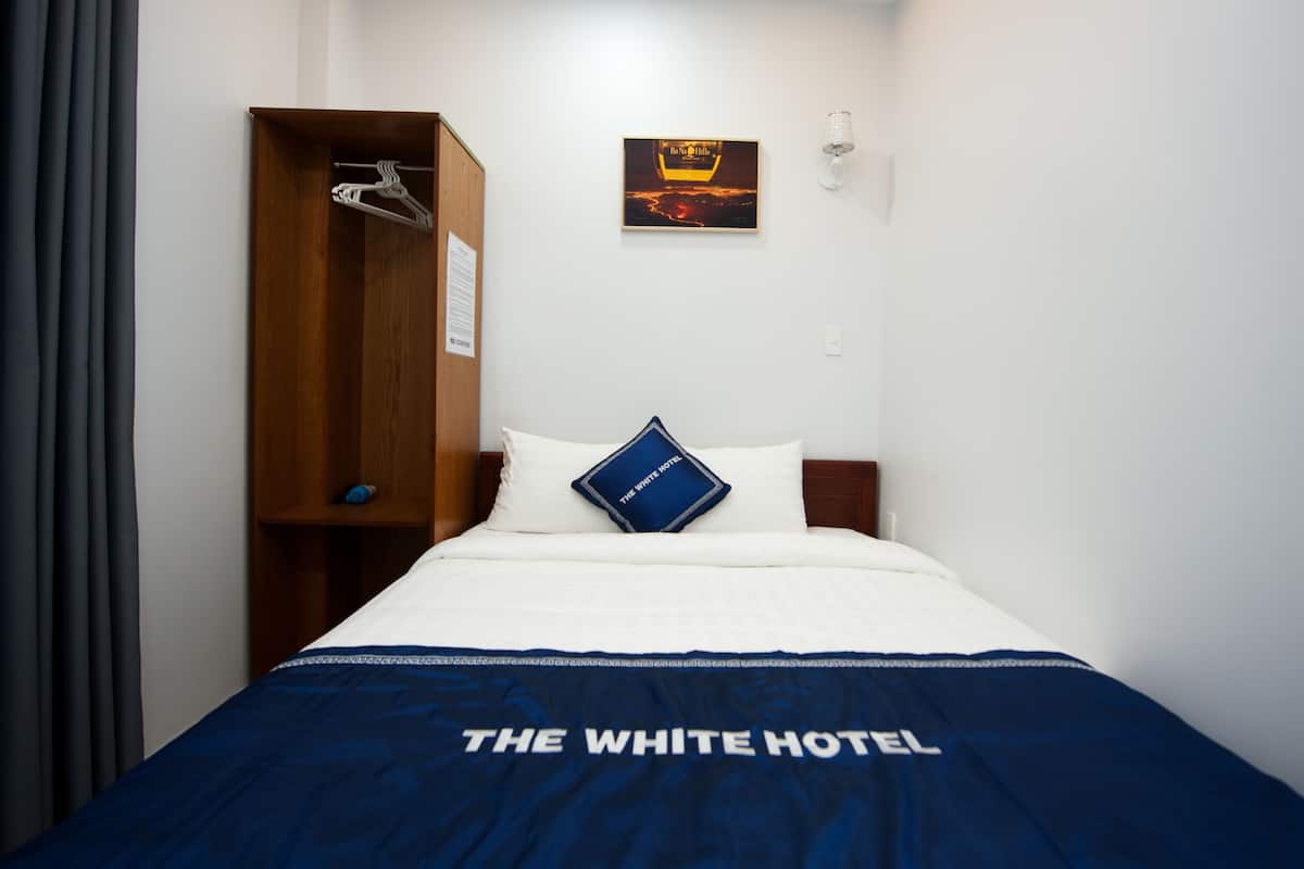 Une chambre d’hôtel comprenant un lit, une armoire, un oreiller orné du logo de « The White Hotel », un tableau accroché au mur et un luminaire.