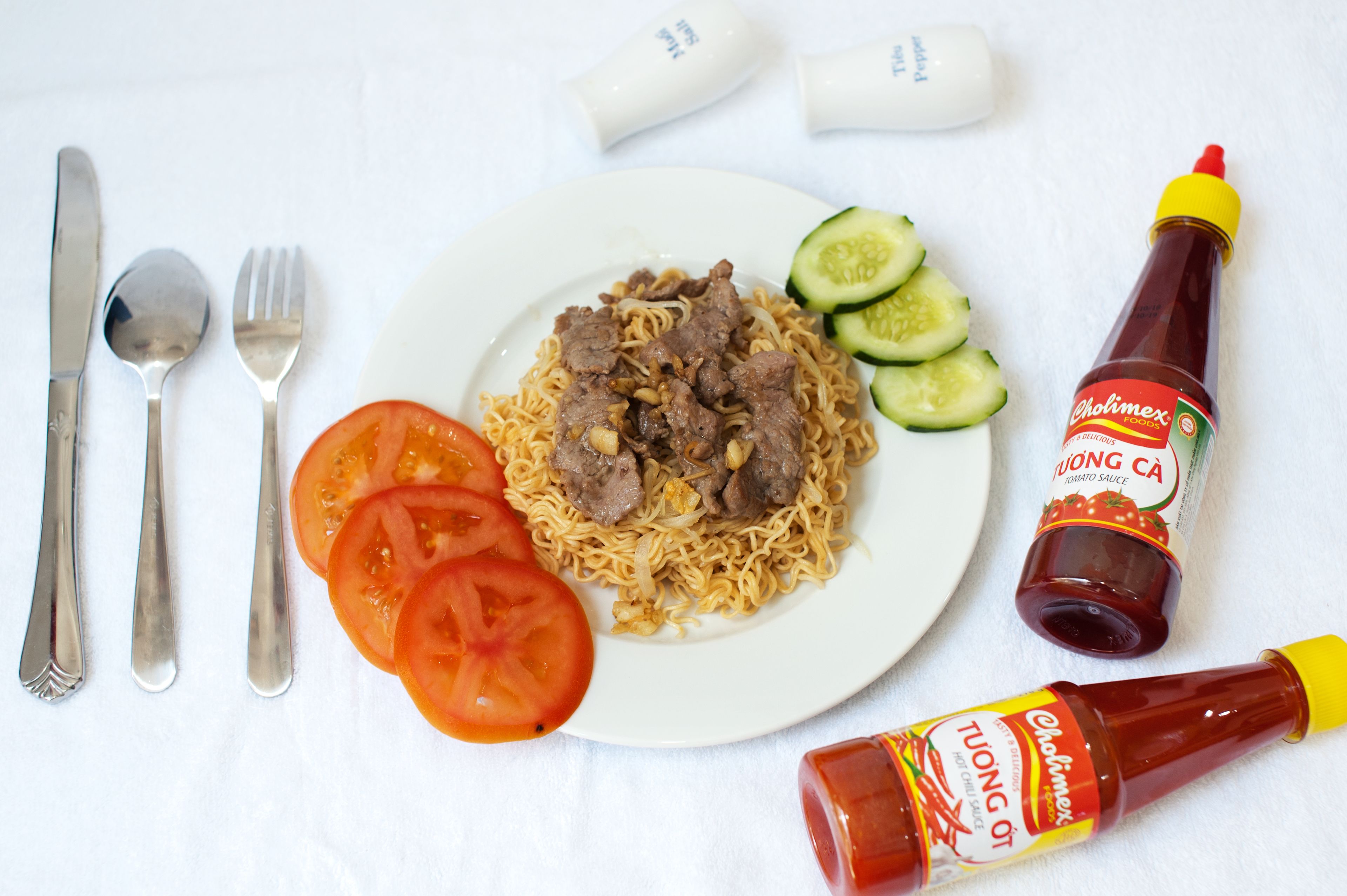 A plate of noodles with meat, cucumber slices, and tomato slices, accompanied by condiments and cutlery on a white surface.