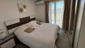 Un lit bien fait, avec une couette blanche et deux oreillers, une table de chevet avec une lampe, un climatiseur fixé au mur et une fenêtre avec des rideaux.