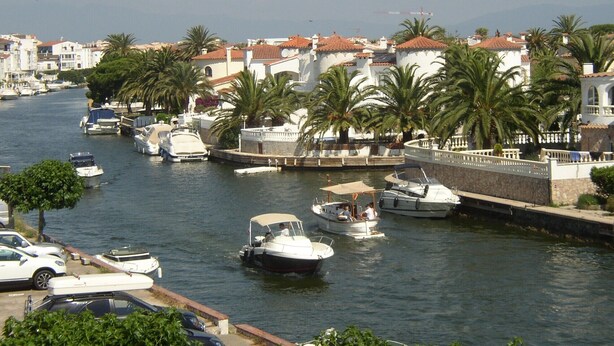 Un port de plaisance où sont amarrés des bateaux, entouré de bâtiments aux toits recouverts de tuiles rouges et de palmiers.