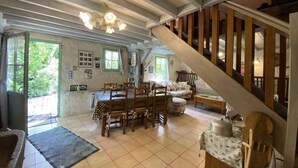 Une salle à manger spacieuse avec un haut plafond et des poutres apparentes, un escalier et une vue sur la verdure environnante.