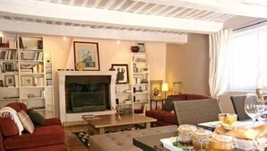A cozy living room with a fireplace, bookshelves, a sofa, and a dining table set with food and drinks. - Les Aiguières en Provence (Suze-la-Rousse)
