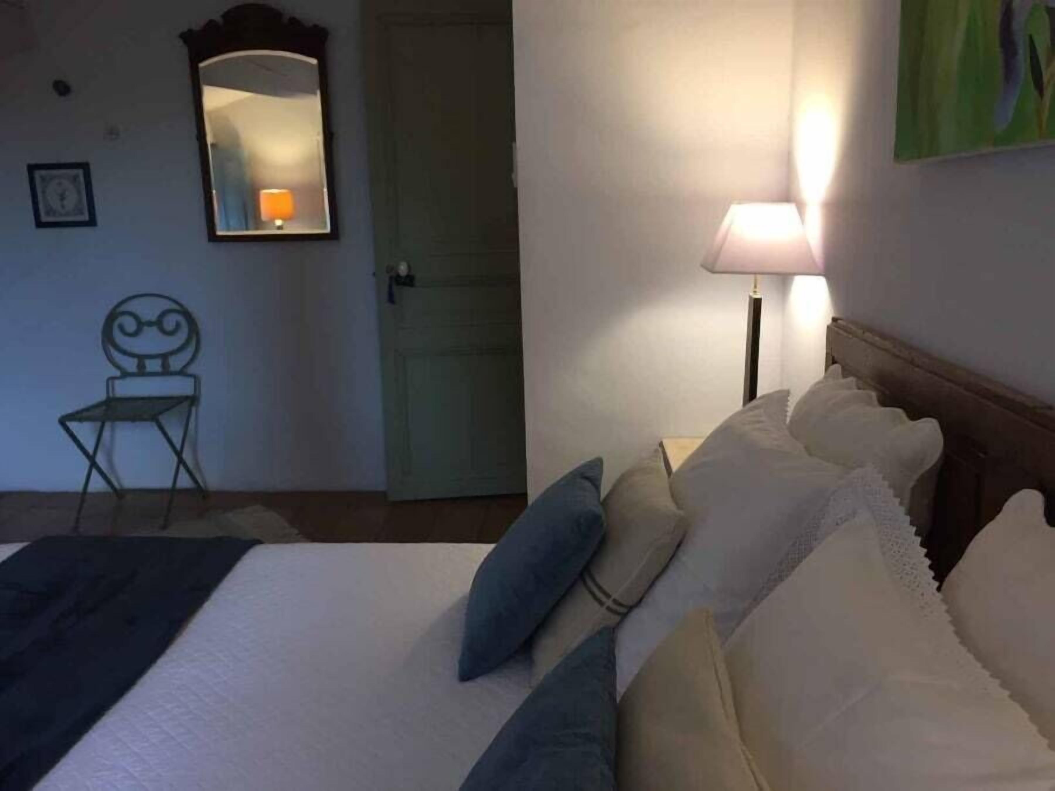 A bedroom with a bed, pillows, a nightstand, a lamp, a chair, a door, and a mirror.