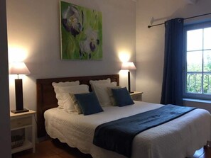 A neatly made bed with a white and blue bedspread, flanked by two bedside tables with lamps, a painting above the bed, and a window with curtains. - Les Aiguières en Provence (Suze-la-Rousse)