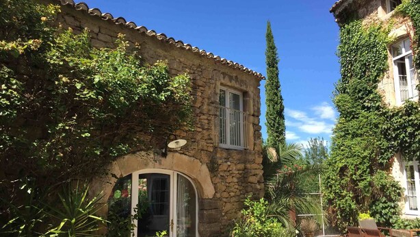A stone building with a tiled roof, a glass door, and a small patio area with a table and chairs. - Les Aiguières en Provence (Suze-la-Rousse)