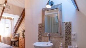 Un baño con un lavamanos blanco, un espejo y un salpicadero de azulejos. Se observa una silla de madera y una cesta de mimbre al fondo.