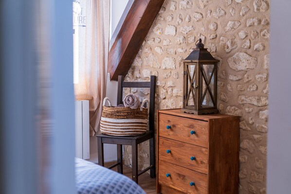 A bedroom with a stone wall, a wooden dresser, a chair with a woven basket, and a window with curtains.