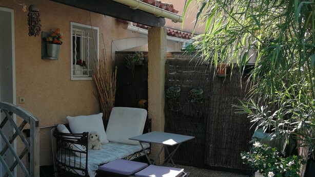 En lille terrasse med en sofa, et bord og en stol. Der er et lille haveområde med en plante og en træbænk.
