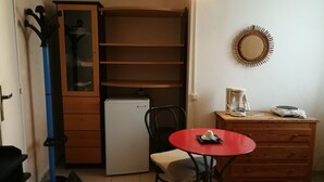 Et kompakt værelse med et lille køleskab, et træskab med glasdøre, et rødt bord med en stol, en kaffemaskine og et spejl på væggen.