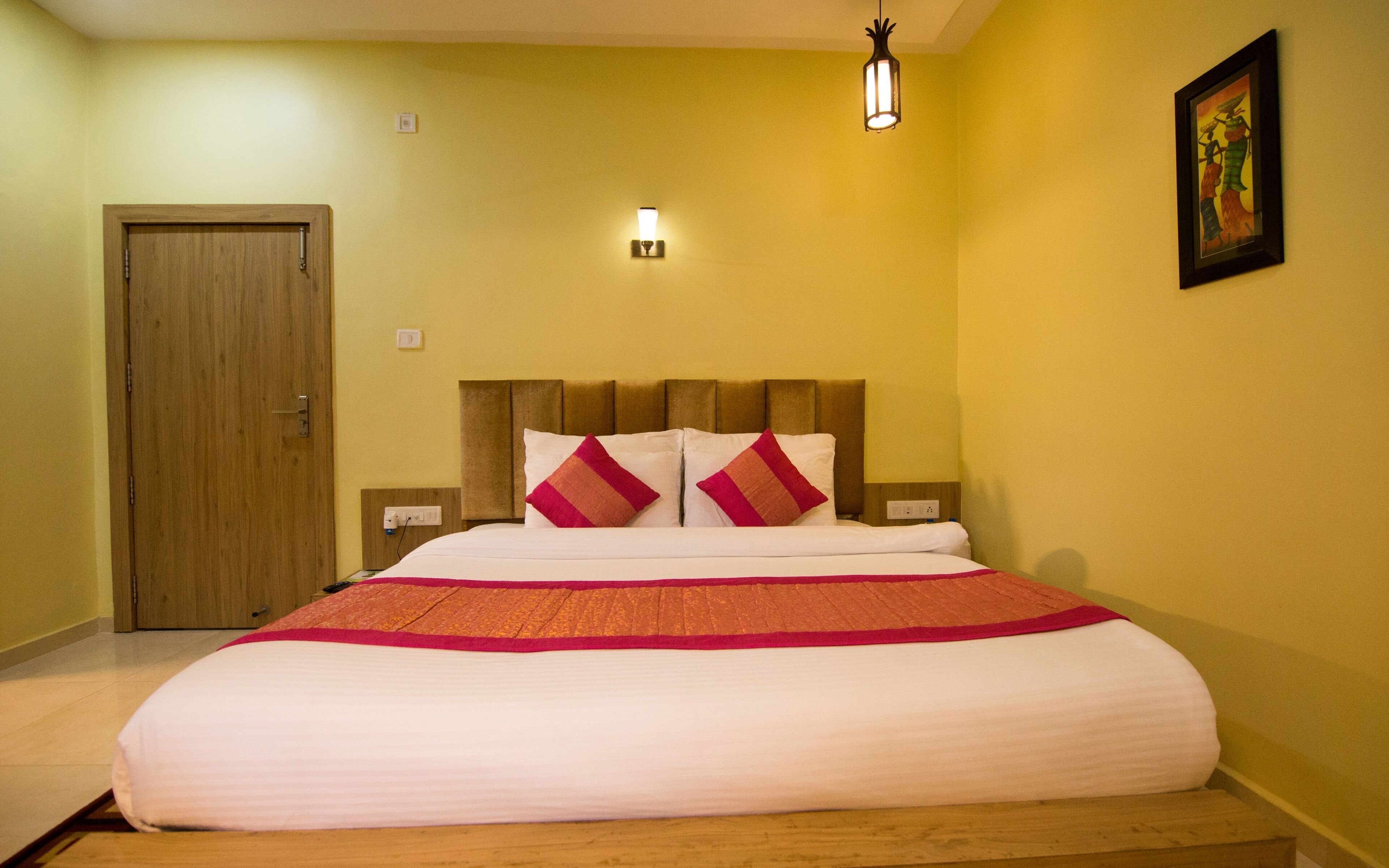 A hotel room with a wooden door, a bed with white bedding and two red pillows, a wall-mounted light, and a framed picture on the wall.