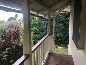 A covered wooden deck with a metal railing, surrounded by lush greenery and a variety of plants. - Wainaku Camp Home (Hilo)