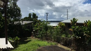 Vista desde debajo de un techo, mostrando un área verde con un sendero pavimentado, rodeado de follaje y un edificio con techo metálico.