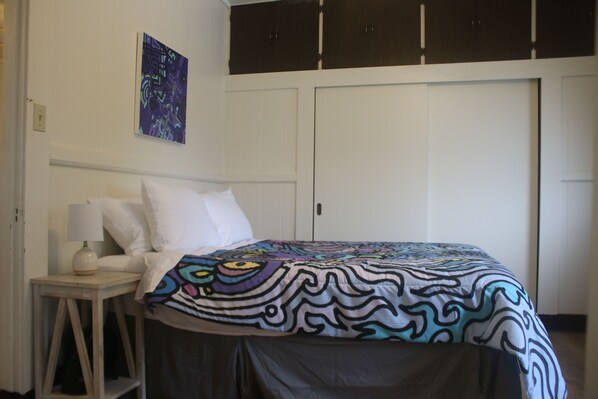A bedroom with a bed, a nightstand, a lamp, a wall art piece, and a wardrobe.