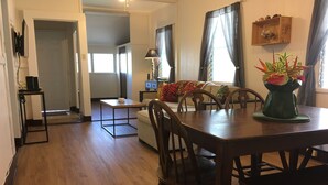 Amplia sala de estar con mesa de comedor de madera, sillas, sofá y mesa de centro. Hay un televisor montado en la pared, una lámpara y una planta en el alféizar de la ventana.