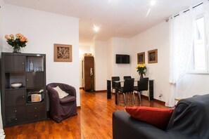A living room with a sofa, a dark cabinet, a dining area with chairs, a TV mounted on the wall, and a window with curtains.