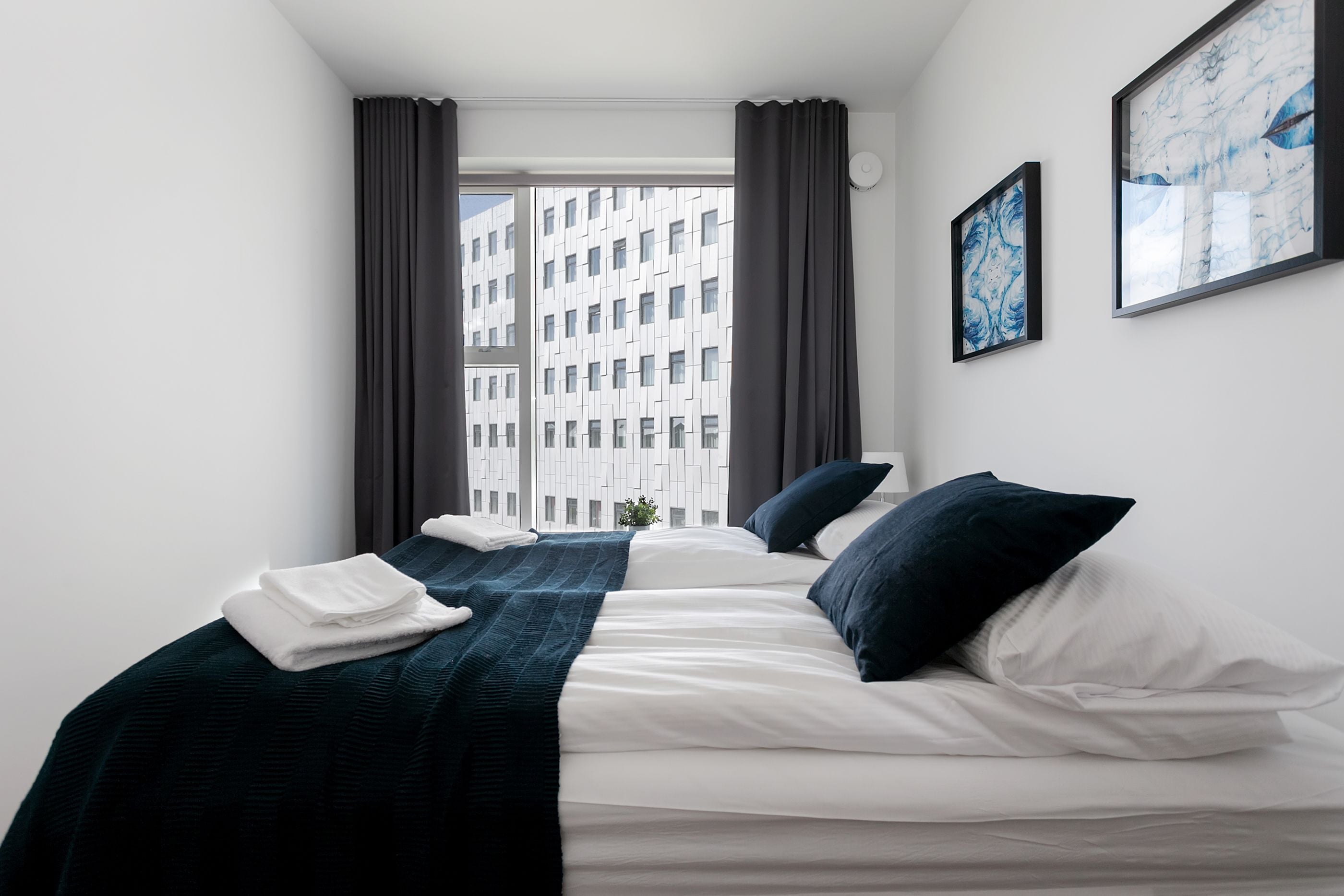 Habitación de hotel con un ventanal grande, cama con ropa de cama blanca y azul oscuro, y dos cuadros en la pared.