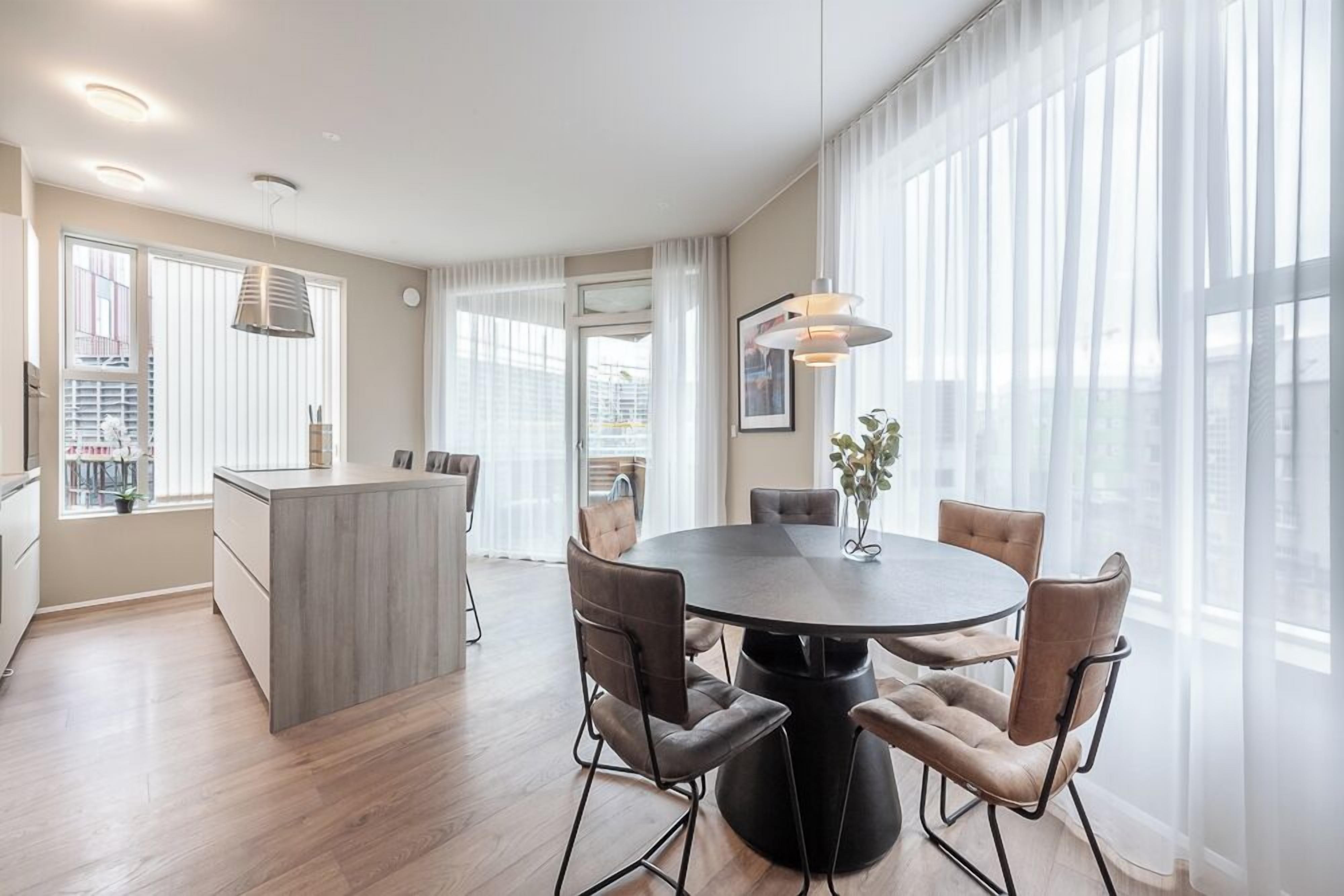 Un espace repas moderne avec une table ronde et six chaises, un coin cuisine doté d’un îlot central et de grandes fenêtres ornées de rideaux légers.