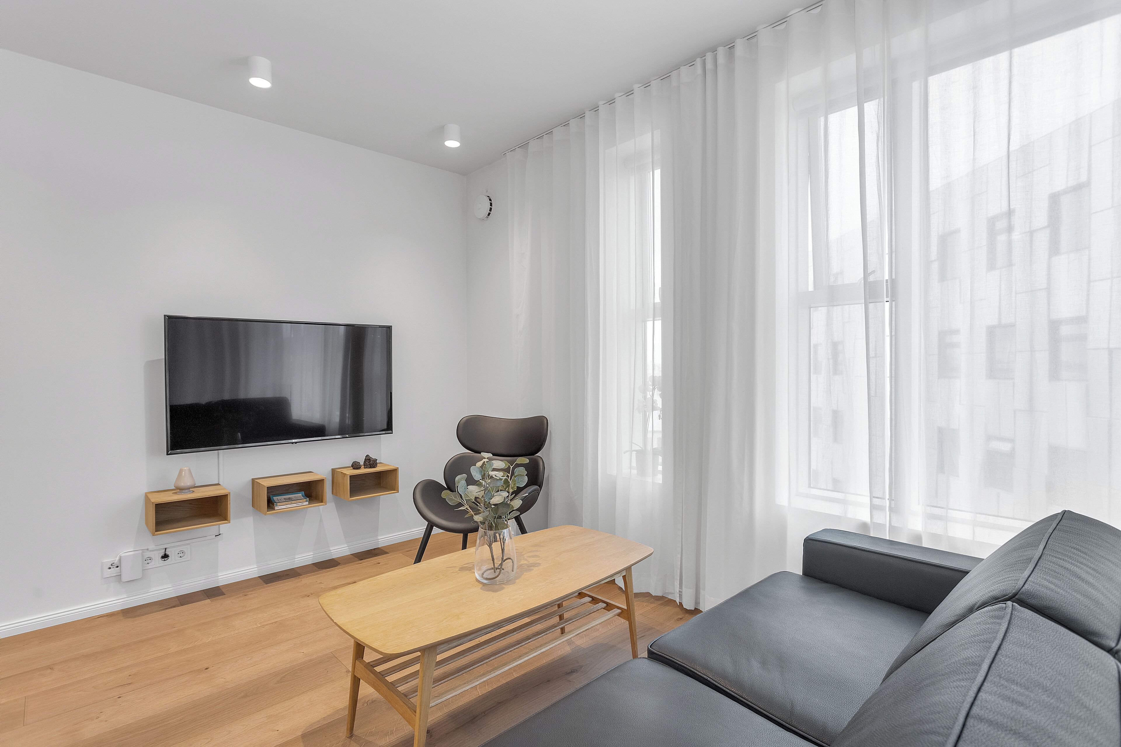 Una sala de estar moderna con un televisor de pantalla plana, una mesa de centro de madera con un florero, un sofá gris y cortinas traslúcidas.