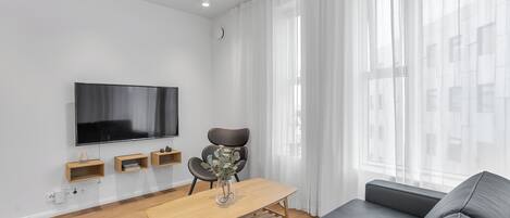 Una sala de estar moderna con un televisor de pantalla plana, una mesa de centro de madera con un florero, un sofá gris y cortinas traslúcidas.