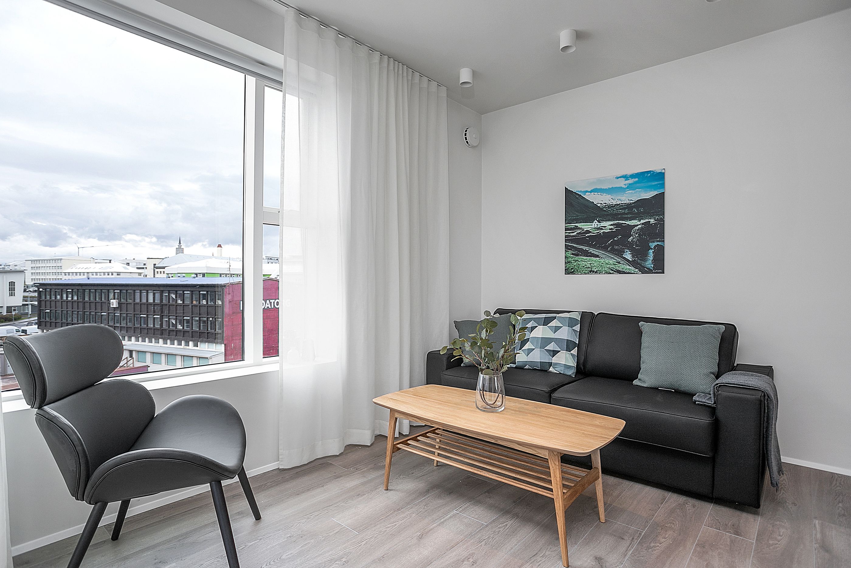 Una sala moderna con un sofá negro, una mesa de centro de madera y una silla negra. Una ventana con cortinas blancas ofrece vistas a edificios.