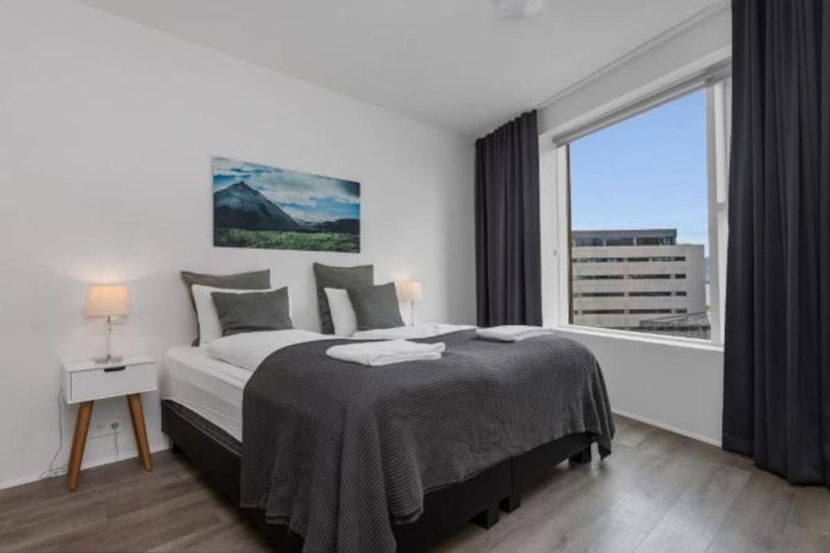 A modern hotel room with a large bed, a nightstand, a window with curtains, and a cityscape view.