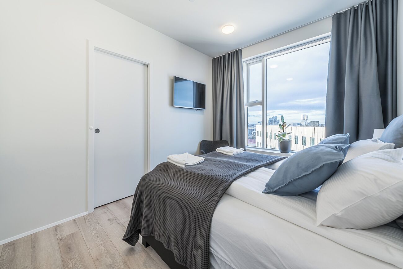 Habitació d'hotel moderna amb un gran finestral, televisió de pantalla plana, llit amb roba de llit blanca i gris, i vista a la ciutat.