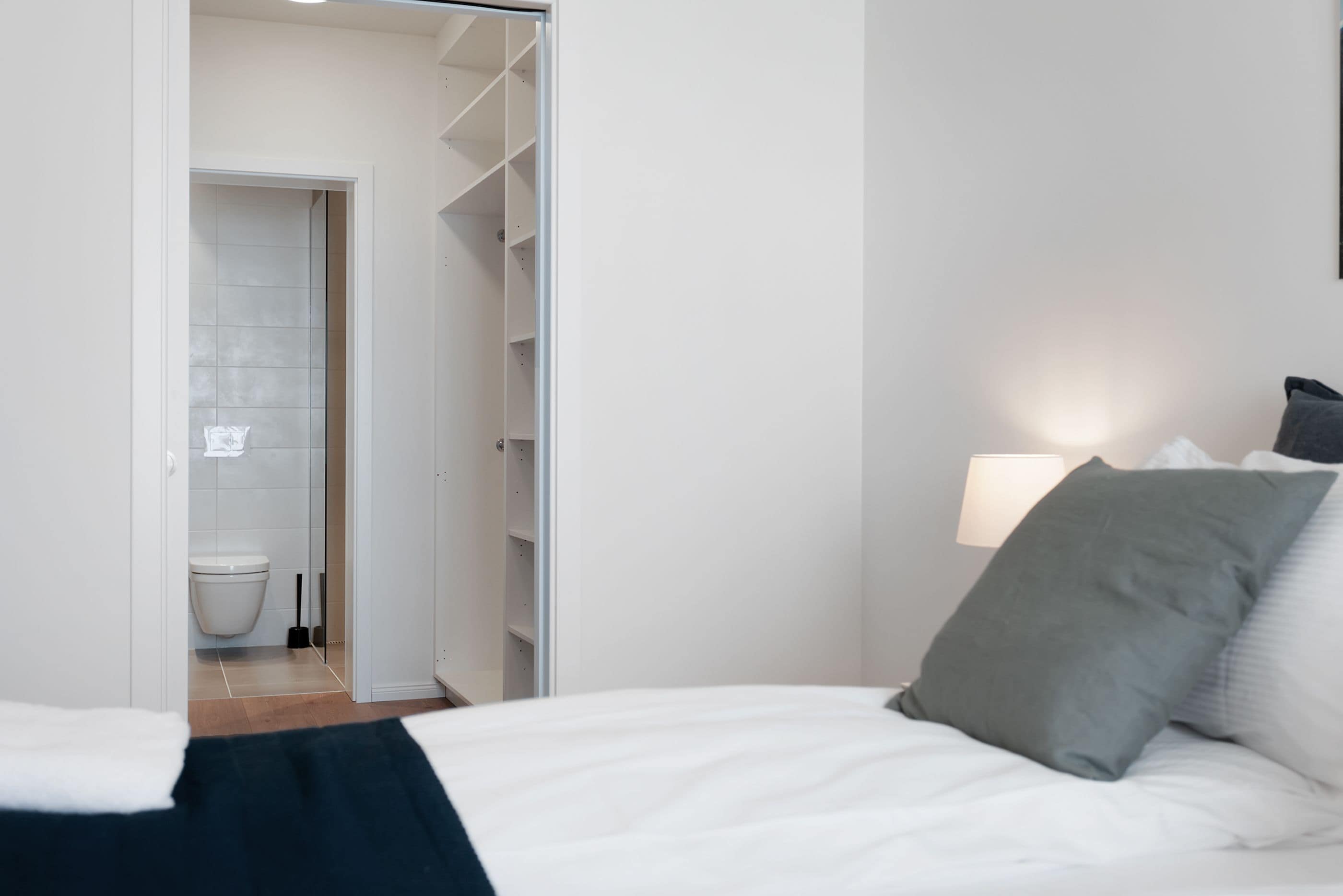 A bedroom with a bed, a nightstand, a wall-mounted lamp, and an open closet leading to a bathroom with a toilet and a broom.