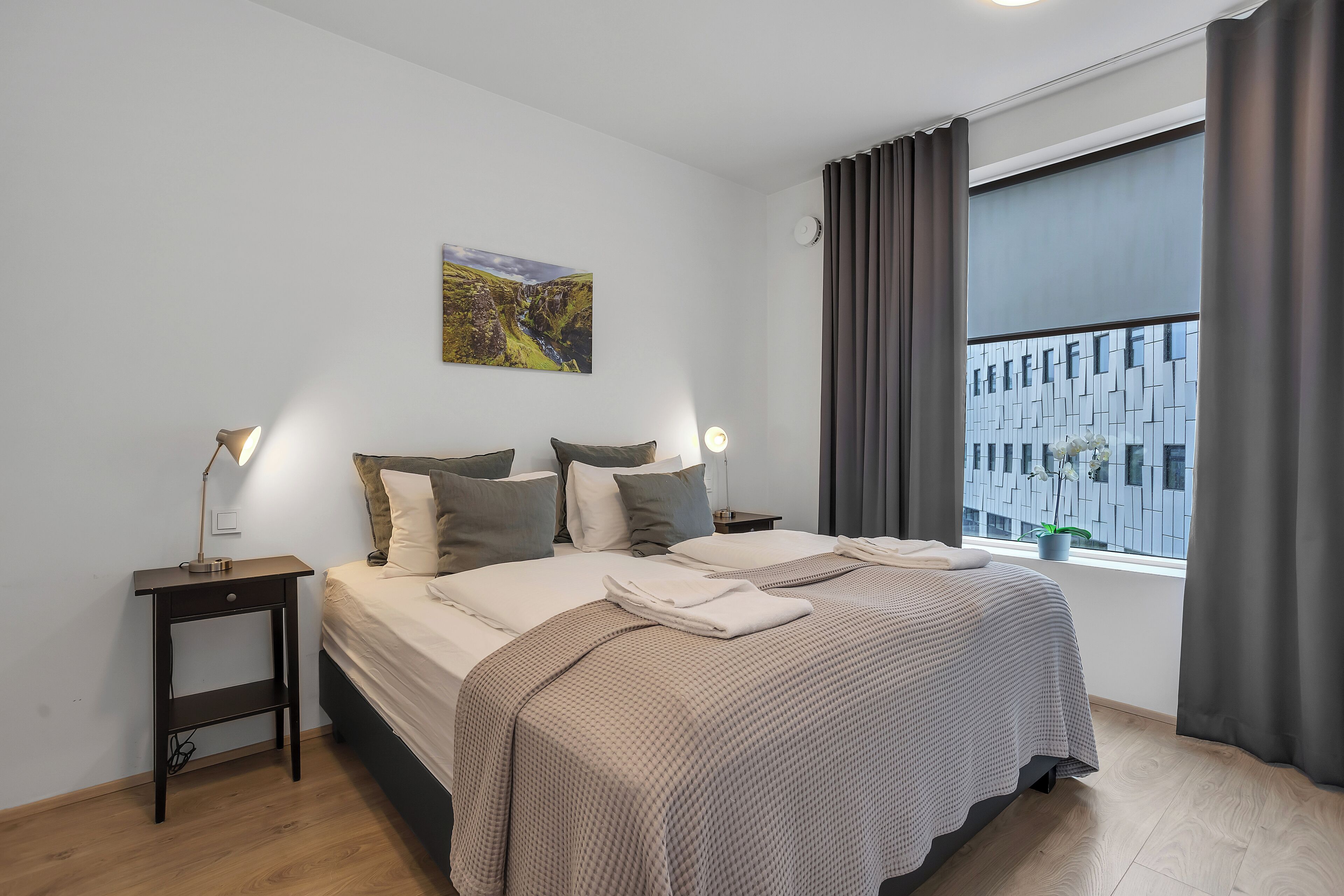 A bedroom with a large bed, two bedside tables, a nightstand, a window with curtains, and a wall-mounted picture.
