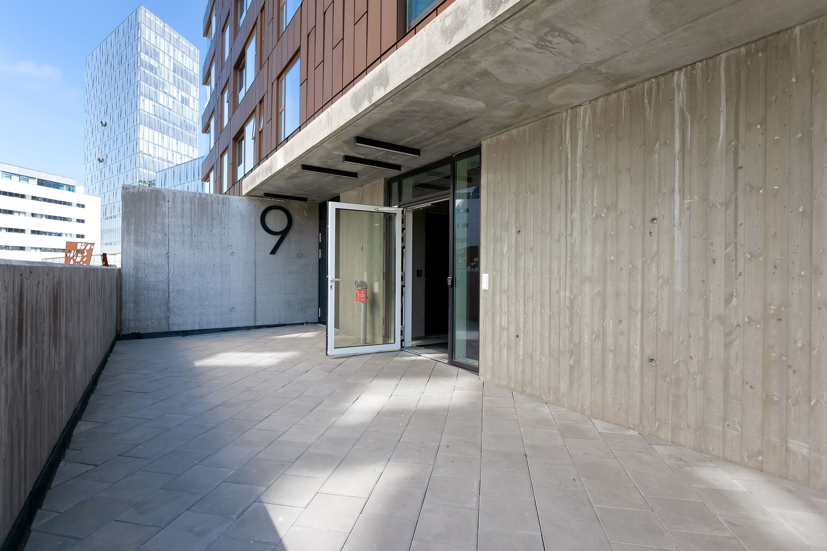 Un espace extérieur doté d’un sol en béton, d’un mur en bois et d’une porte vitrée donnant accès à un bâtiment.