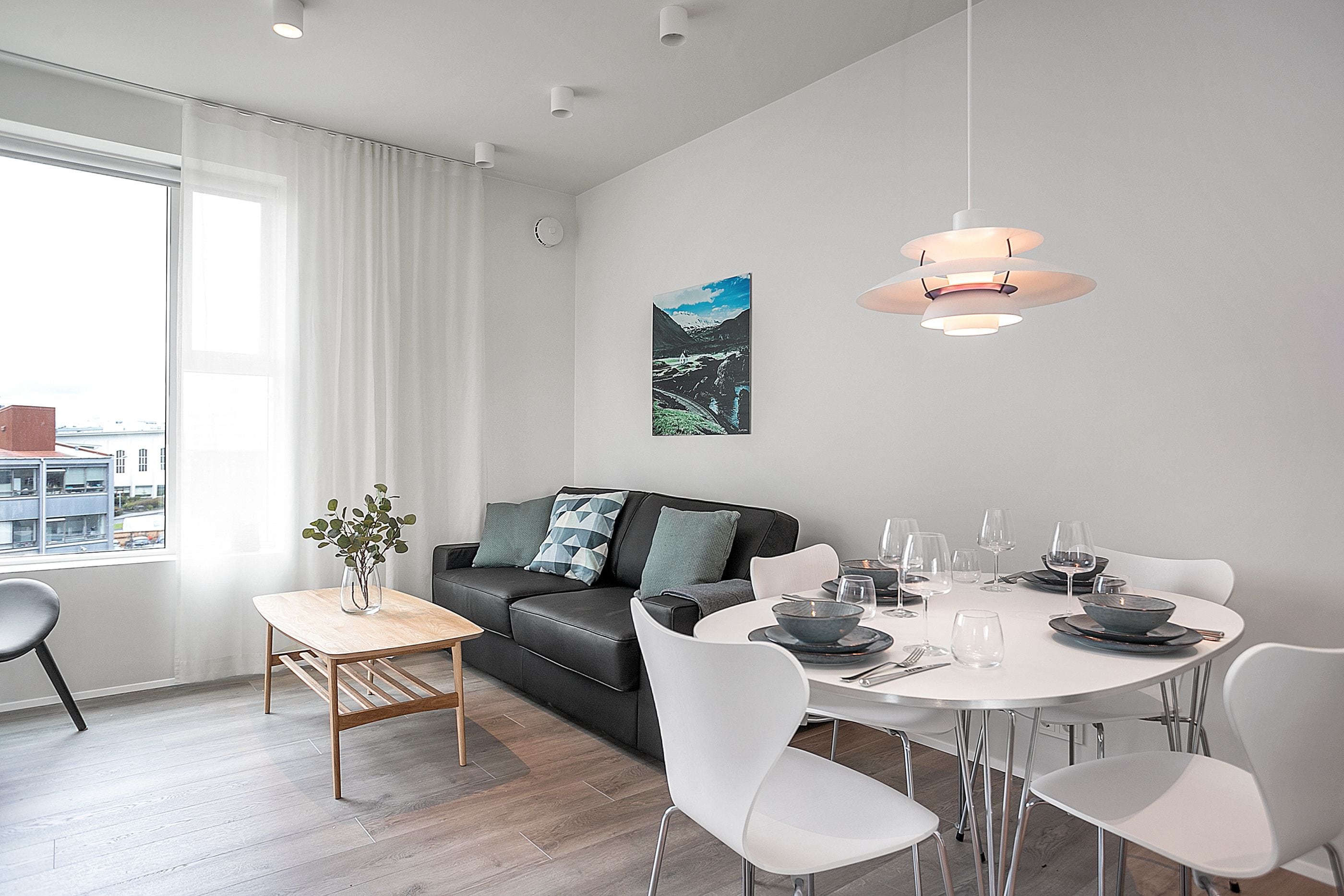 A modern living room with a dining area, featuring a sofa, a dining table set for four, and a large window with curtains.